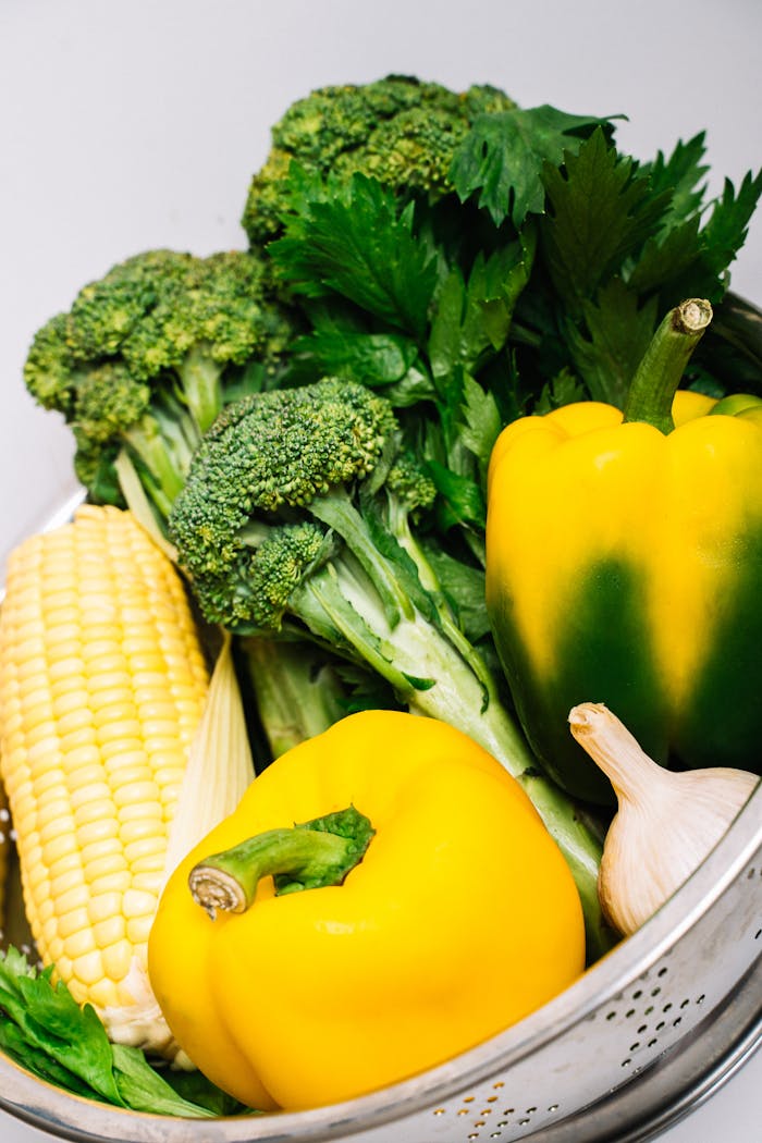 A variety of fresh vegetables including bell peppers, broccoli, and corn in a metal strainer.